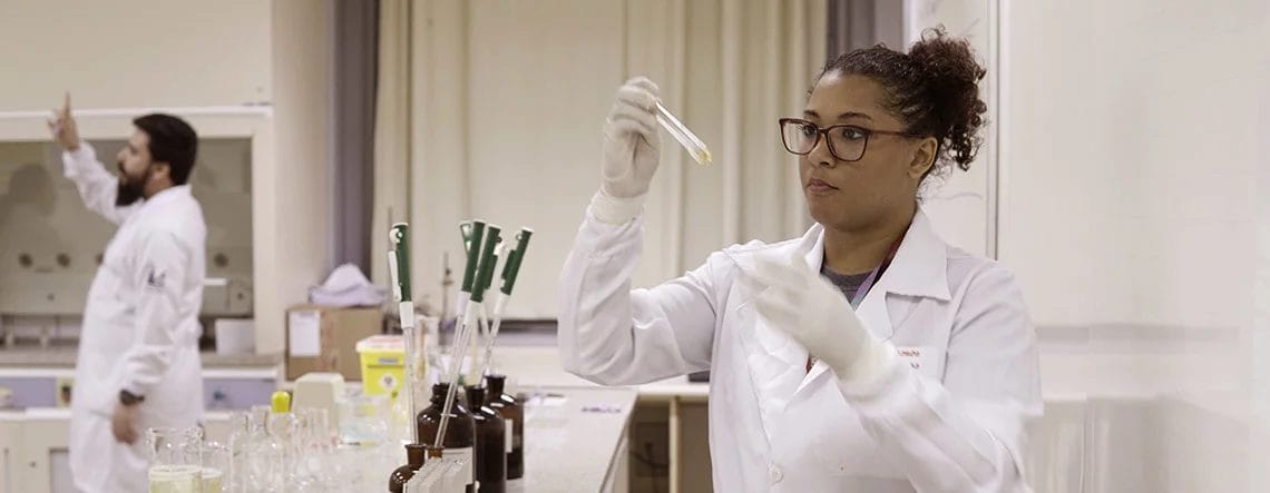 mulher de jaleco em laboratório analisa conteúdo de um frasco