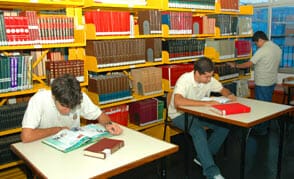 Dois alunos sentados em mesas lendo livros, um terceiro aluno no fundo olha as prateleiras
