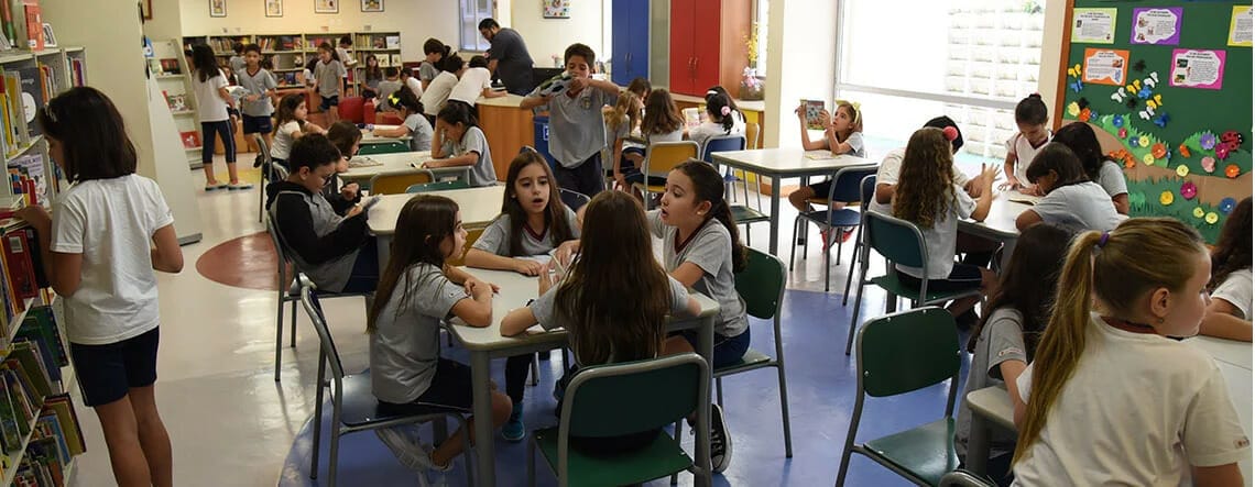 crianças sentadas em grupo em mesas na biblioteca do colégio