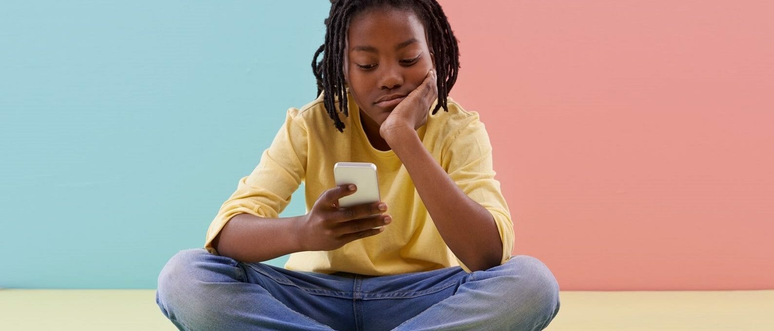 adolescente sentada em frente a um fundo colorido, mexendo no celular com cara emburrada