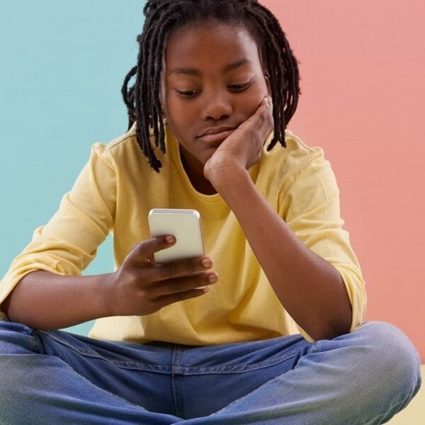 adolescente sentada em frente a um fundo colorido, mexendo no celular com cara emburrada