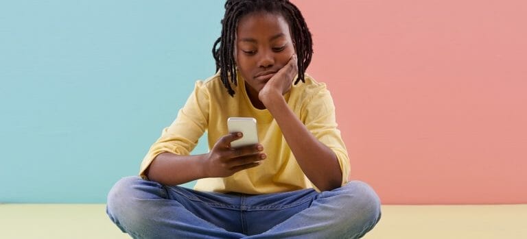 adolescente sentada em frente a um fundo colorido, mexendo no celular com cara emburrada