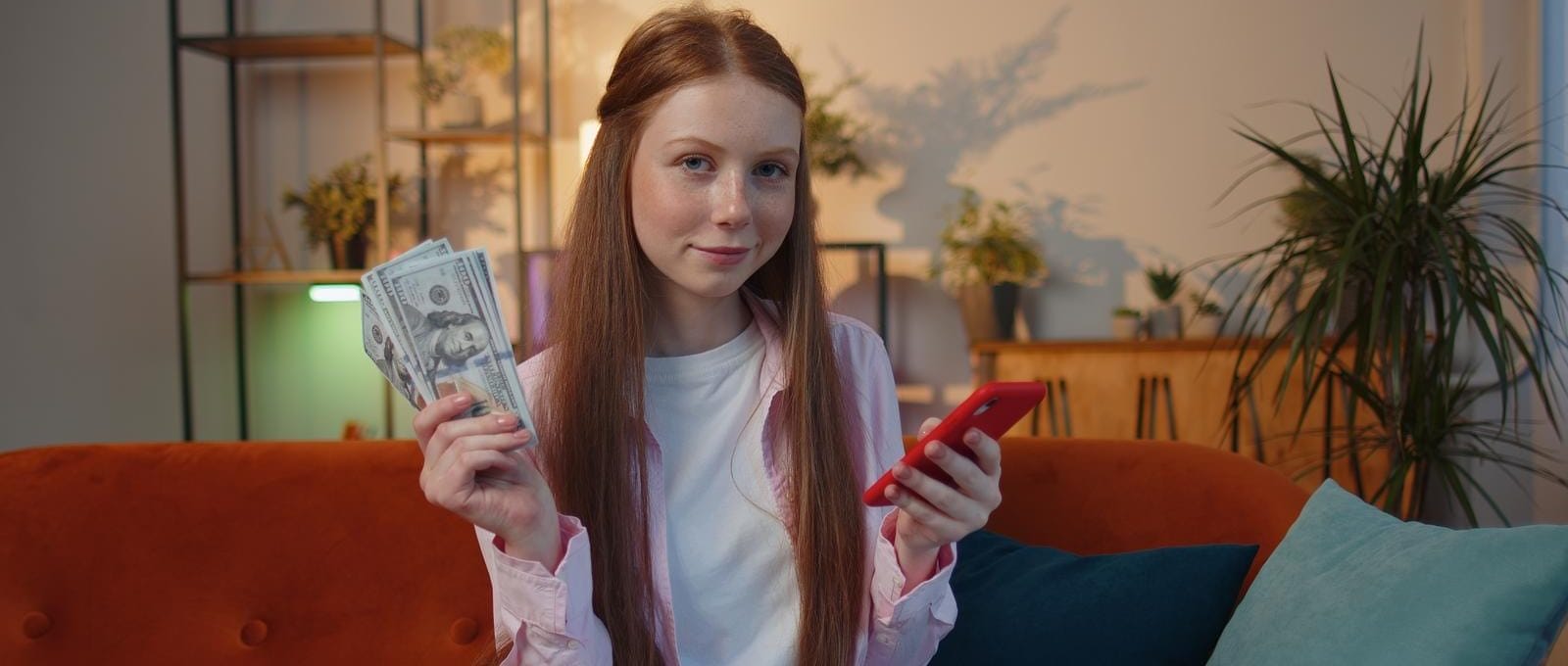 adolescente ruiva sentada no sofá da sala sorrindo, com notas de dinheiro em uma mão, e o celular na outra
