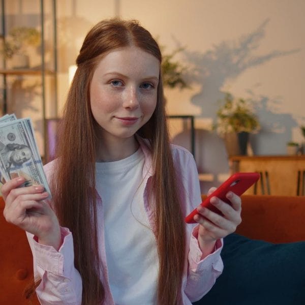adolescente ruiva sentada no sofá da sala sorrindo, com notas de dinheiro em uma mão, e o celular na outra