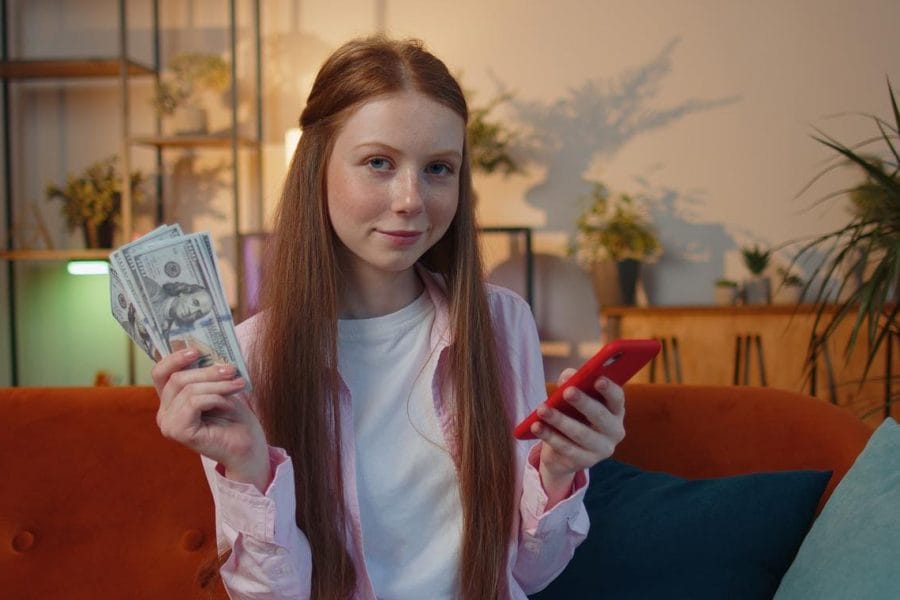 adolescente ruiva sentada no sofá da sala sorrindo, com notas de dinheiro em uma mão, e o celular na outra