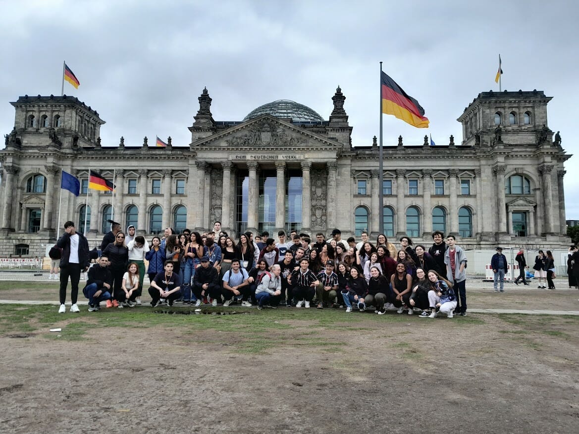 turma de alunos do colégio cruzeiro reunidos para foto em frente a um prédio histórico da alemanha, com bandeiras do país tremulando ao fundo