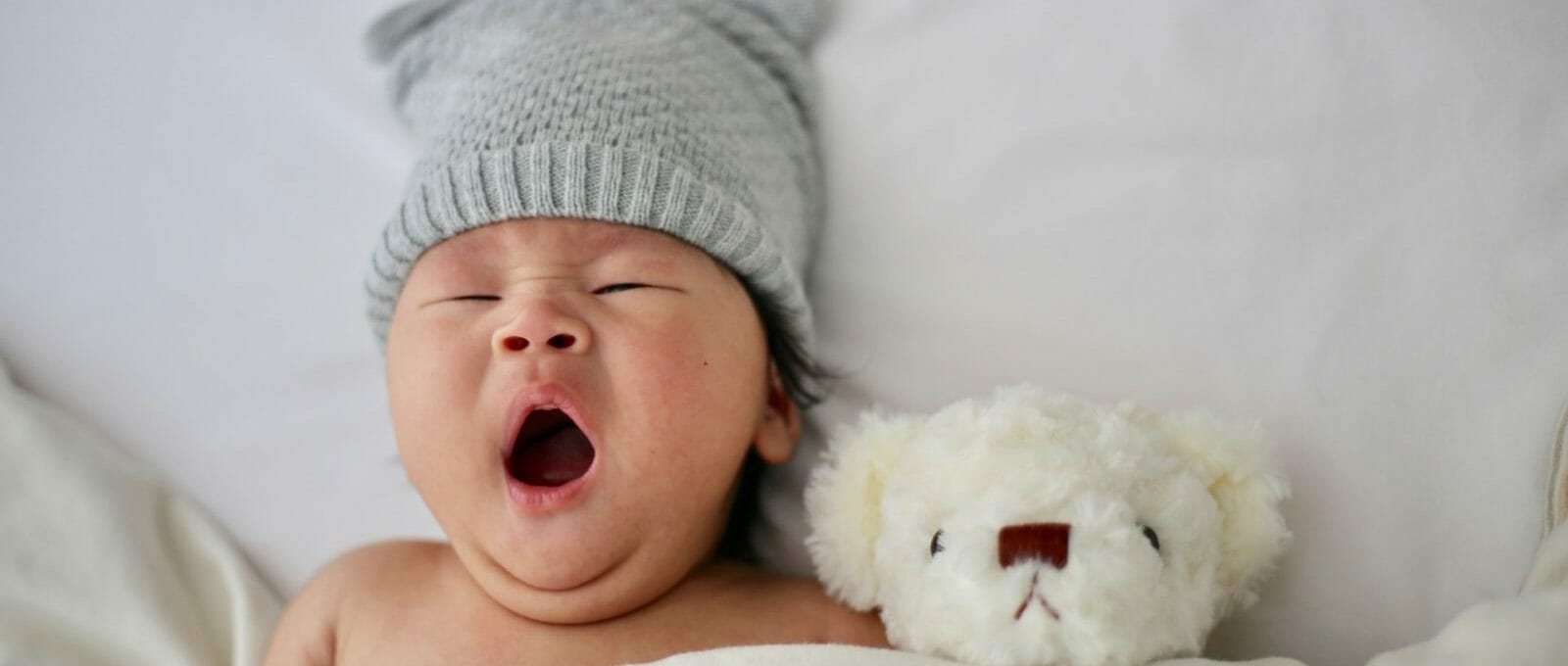 bebê bocejando no berço usando gorrinho, com cobertor e ursinho de pelúcia ao lado