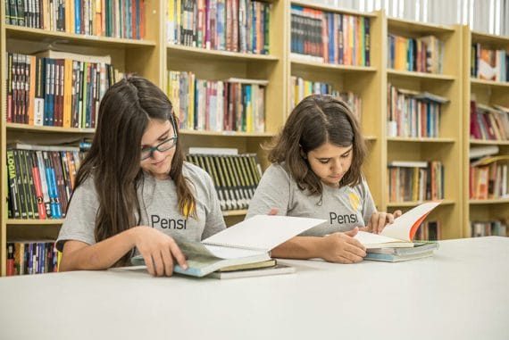 duas alunas lendo em mesa da biblioteca, com estante de livros ao fundo