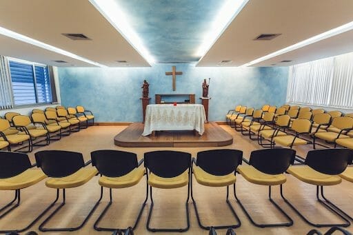 Imagem de uma capela vazia. Ao centro, imagem da cruz, cercada por cadeiras e com luzes brancas iluminando o teto.