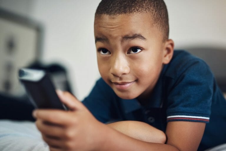 Criança deitada na cama com controle remoto nas mãos, representando o conceito de controlar o próprio tempo de tela