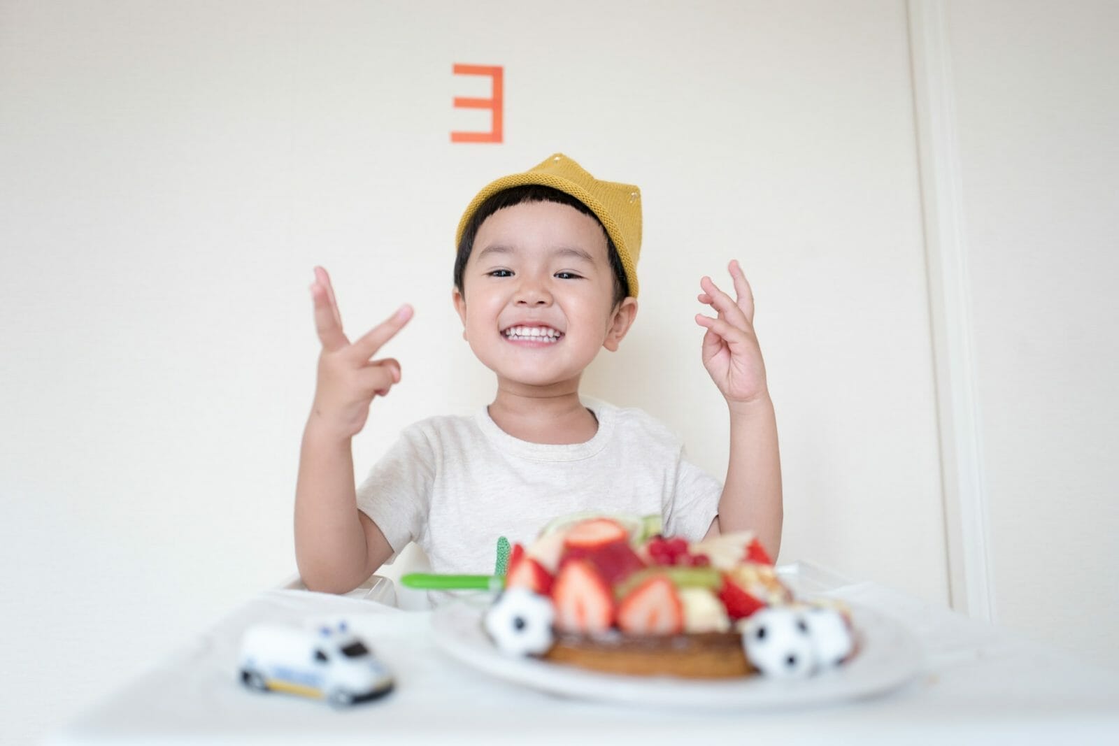 criança oriental sorrindo sentada em uma mesa com um carrinho e um bolo de aniversário