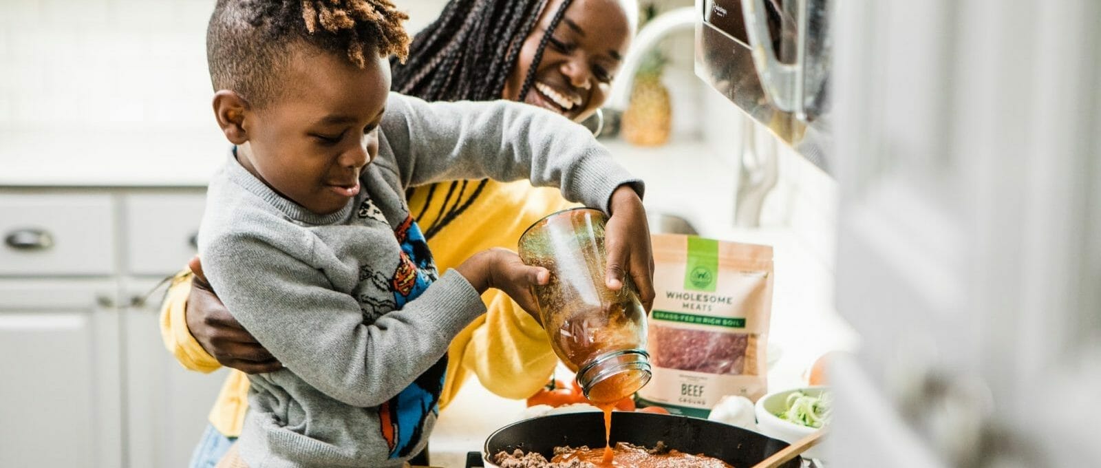 criança colocando molho na carne antes de ir ao fogo, enquanto a mãe sorri ao lado e o ajuda
