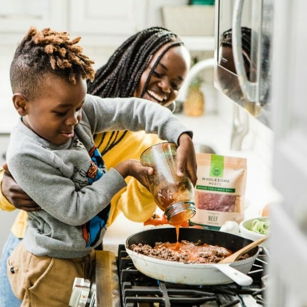 criança colocando molho na carne antes de ir ao fogo, enquanto a mãe sorri ao lado e o ajuda