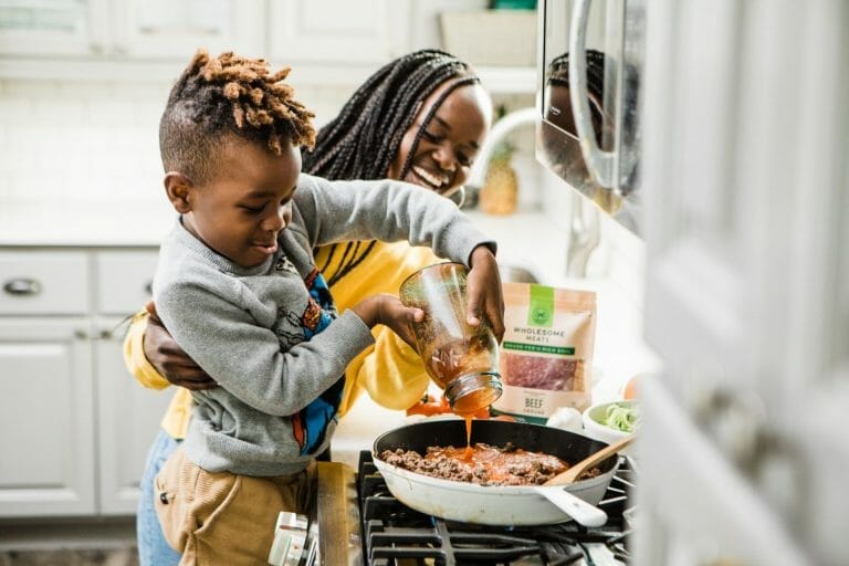 criança colocando molho na carne antes de ir ao fogo, enquanto a mãe sorri ao lado e o ajuda