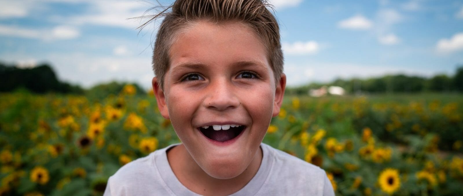 Uma criança loira sorri em primeiro plano. Ao fundo, é possível ver um campo de girassóis desfocado.