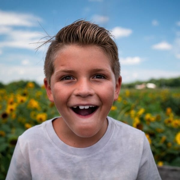 Uma criança loira sorri em primeiro plano. Ao fundo, é possível ver um campo de girassóis desfocado.