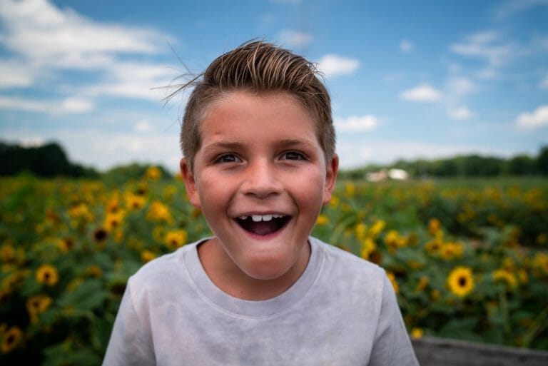 Uma criança loira sorri em primeiro plano. Ao fundo, é possível ver um campo de girassóis desfocado.