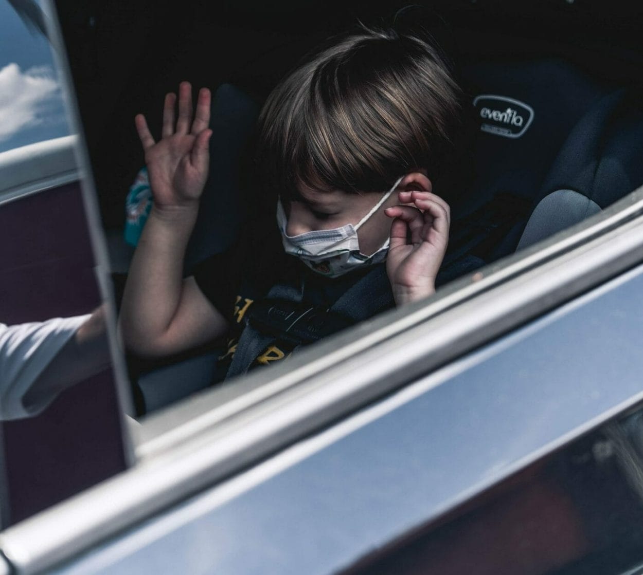 criança de máscara sentada no sozinha no banco de trás do carro com as mãos para cima