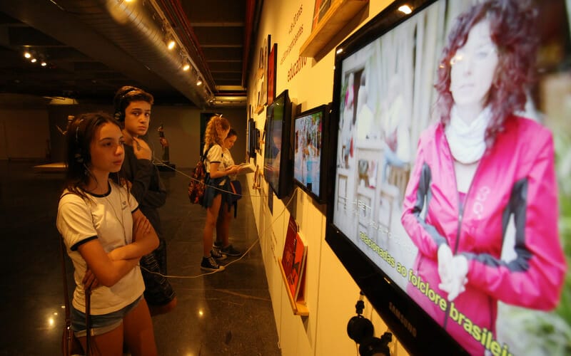 alunos do ensino fundamental visitando exposição em um museu e ouvindo áudios sobre as obras que passam em telas presas à parede