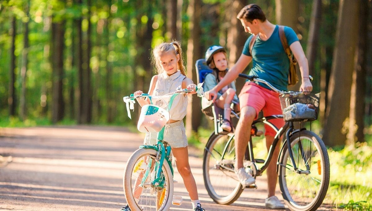 Pai arrumando bebê na cadeirinha da bicicleta e menina andando de bike ao lado dele