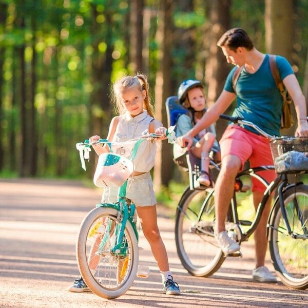 Pai arrumando bebê na cadeirinha da bicicleta e menina andando de bike ao lado dele