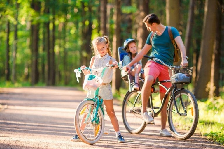 Pai arrumando bebê na cadeirinha da bicicleta e menina andando de bike ao lado dele