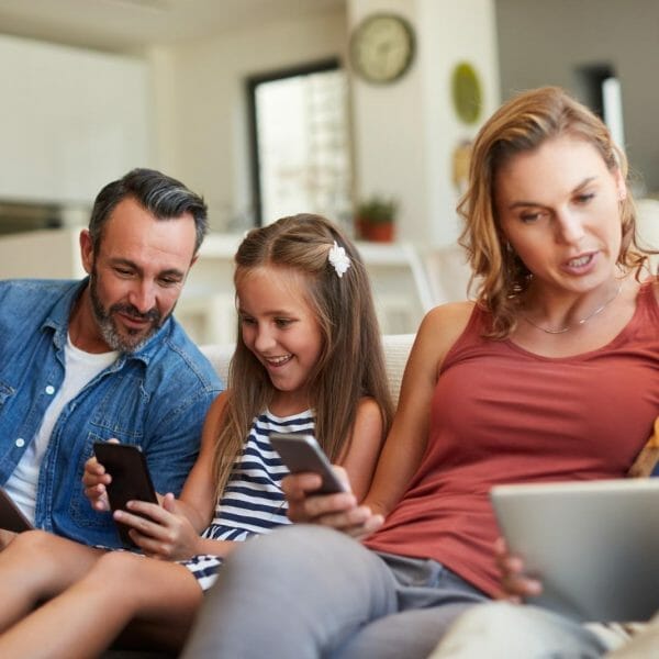 pai, filha, mãe e filho sentados no sofá olhando para celulares, tablet e notebook