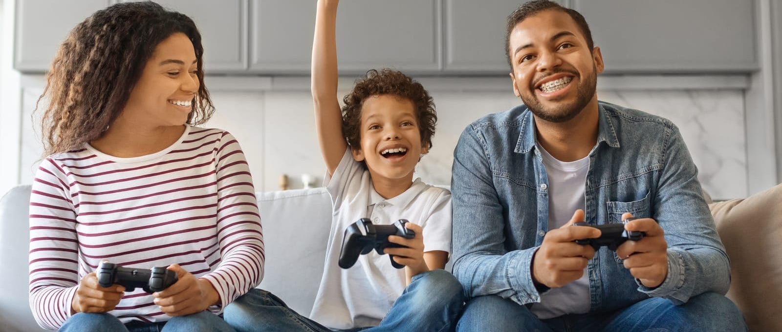 Família sentada no sofá, comemorando enquanto joga videogame, sorrindo e segurando os controles olhando para a televisão