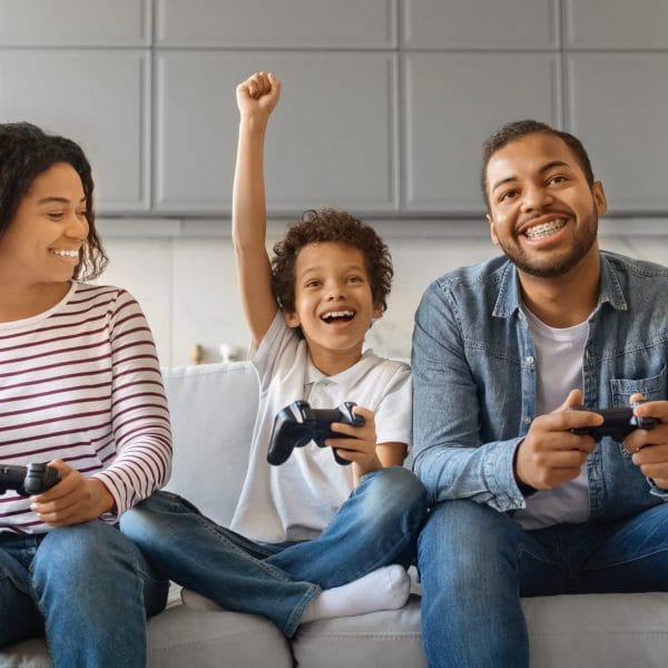 Família sentada no sofá, comemorando enquanto joga videogame, sorrindo e segurando os controles olhando para a televisão