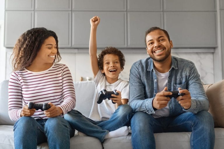 Família sentada no sofá, comemorando enquanto joga videogame, sorrindo e segurando os controles olhando para a televisão