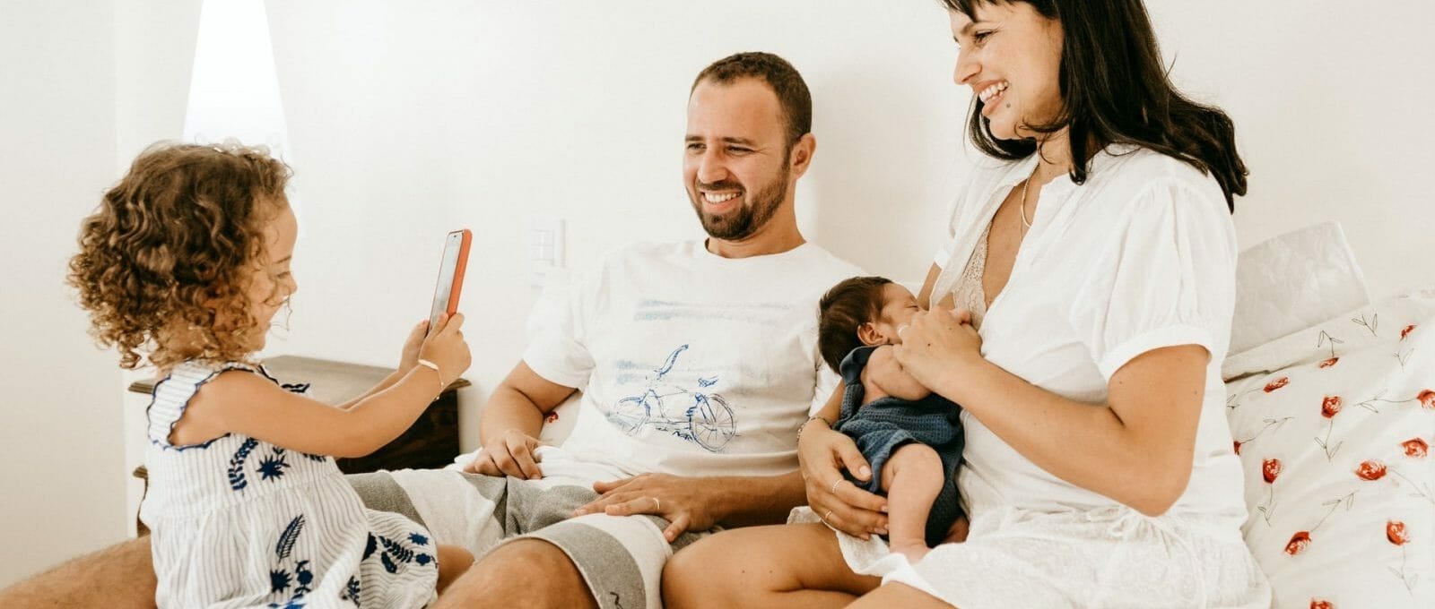família passando tempo junta, dando de mamar à filha bebê enquanto a filha criança fotografa os pais com o celular