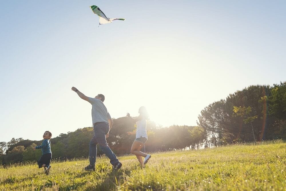 pai soltando pipa com os dois filhos em um campo aberto, em um dia de sol