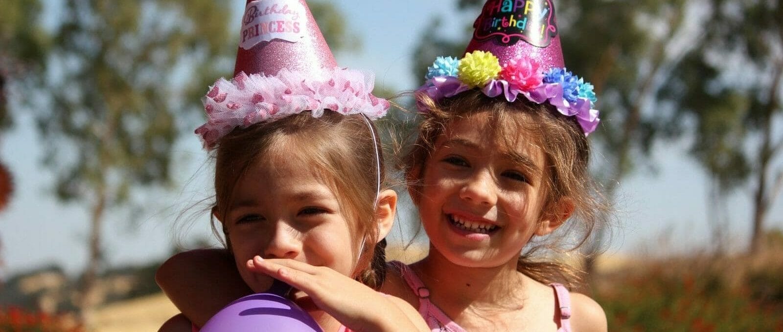 Duas meninas comemorando o aniversário. Ambas estão com chapéus de festa coloridos e uma assopra um balão roxo