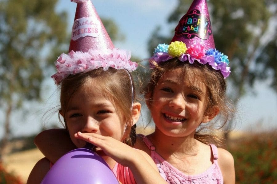 Duas meninas comemorando o aniversário. Ambas estão com chapéus de festa coloridos e uma assopra um balão roxo