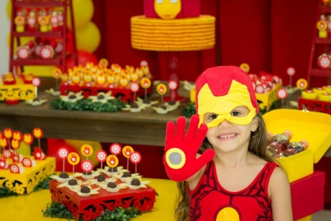 menina usando vestido vermelho, luva e máscara do homem de ferro. ao fundo, mesa de doces decorada em vermelho e amarelo, cores do herói