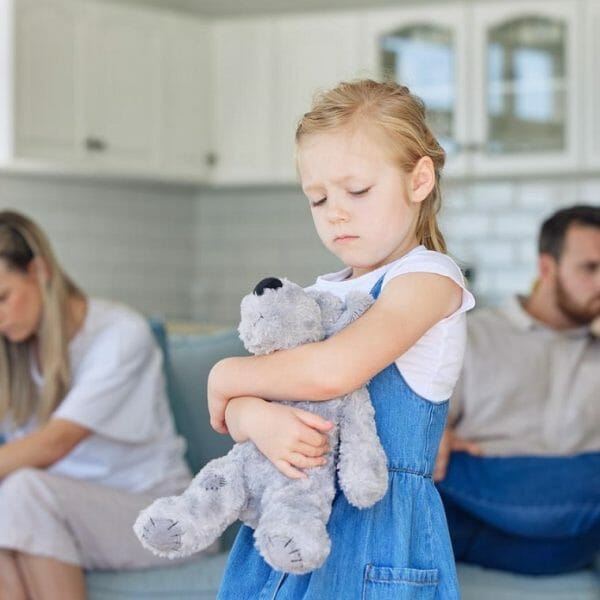 pais brigados ao fundo, com criança em primeiro plano, triste e abraçando um ursinho, representando o conceito de criar filho com pais separados