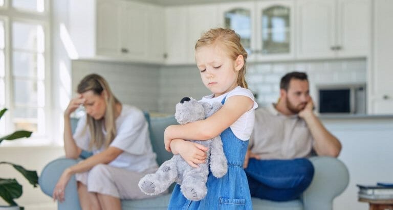 pais brigados ao fundo, com criança em primeiro plano, triste e abraçando um ursinho, representando o conceito de criar filho com pais separados