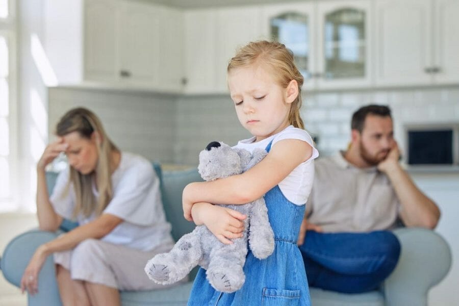 pais brigados ao fundo, com criança em primeiro plano, triste e abraçando um ursinho, representando o conceito de criar filho com pais separados