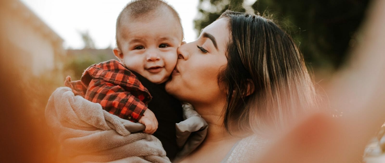 mãe dando beijo em bebê e segurando no colo ao ar livre