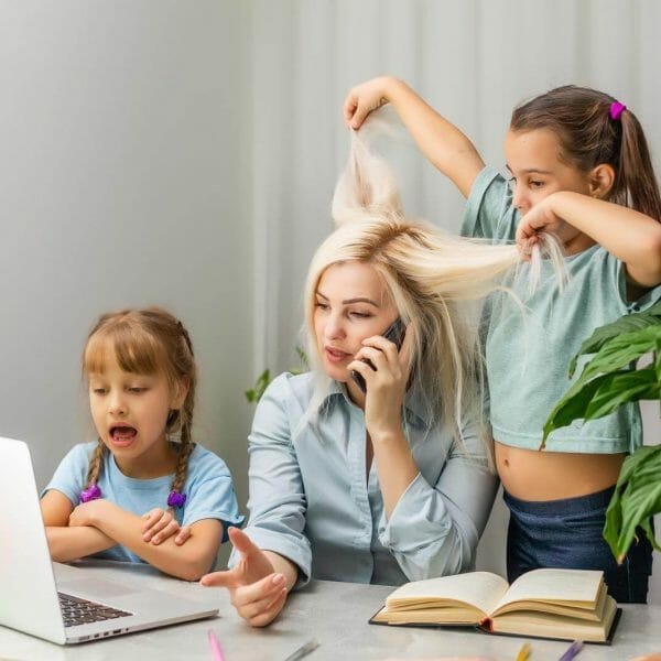 mãe usando o computador e falando ao telefone enquanto uma filha puxa os seus cabelos e a outra fala ao seu lado, representando os desafios da maternidade no mercado de trabalho
