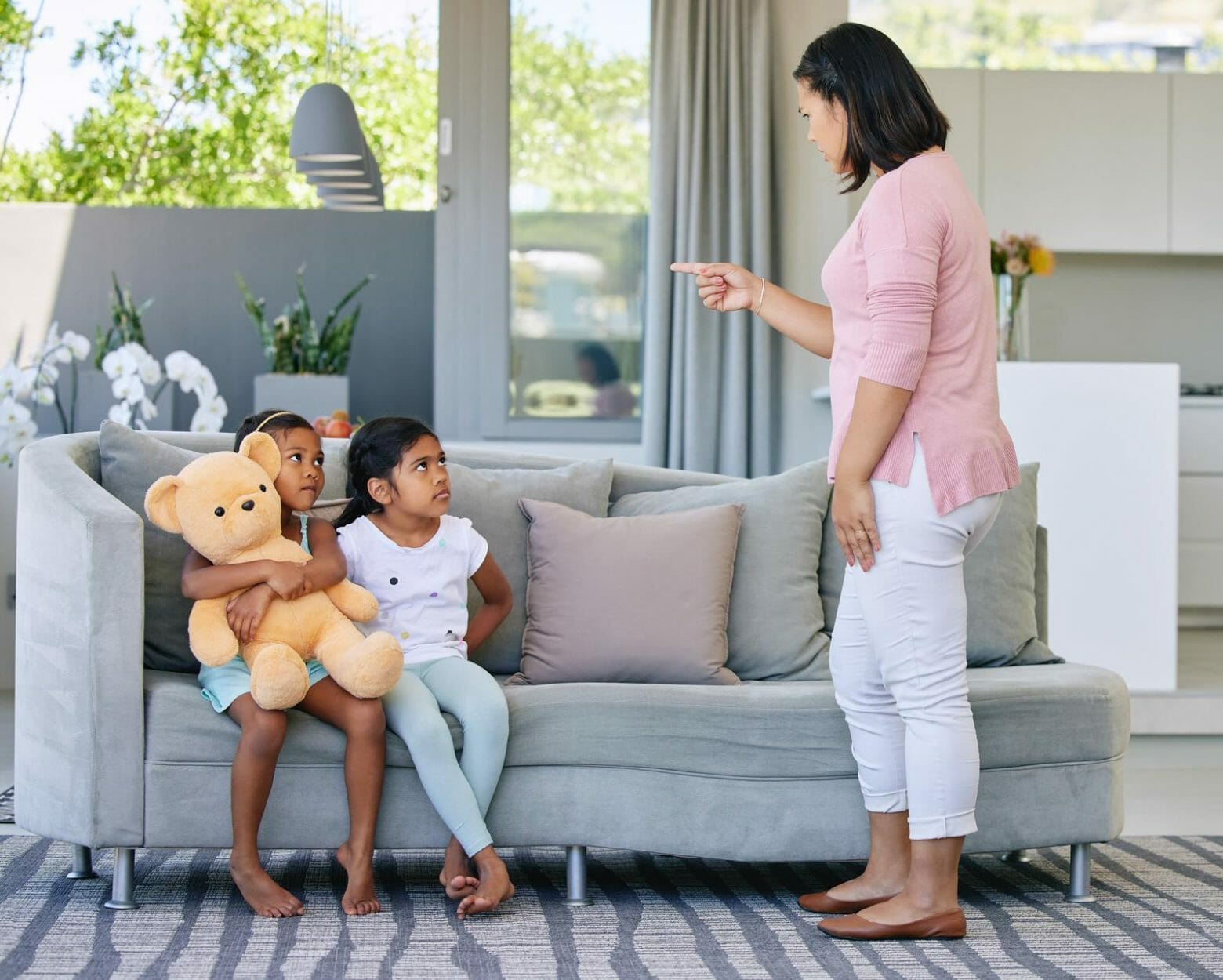 Duas meninas sentadas no sofá, uma delas segurando um urso de pelúcia grande, ouvindo a mãe dando uma bronca na sala 