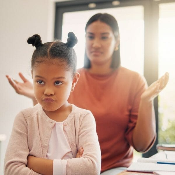 mãe atrás da filha brigando com ela; a menina olha para a câmera de braços cruzados e cara emburrada, de costas para a mãe