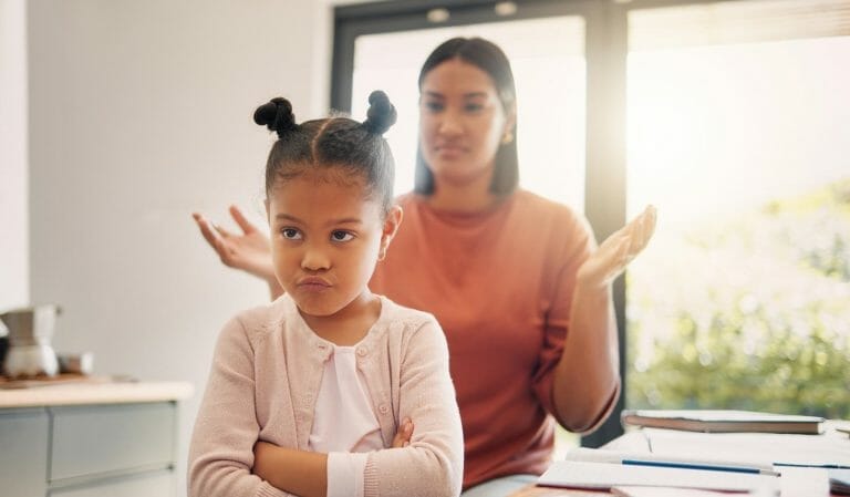 mãe atrás da filha brigando com ela; a menina olha para a câmera de braços cruzados e cara emburrada, de costas para a mãe