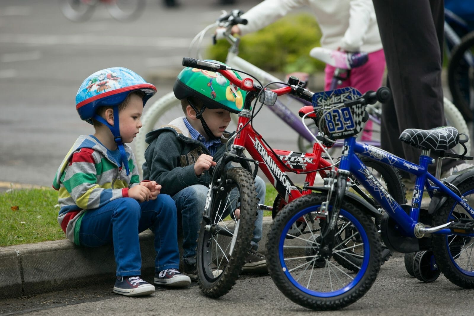 Crianças de capacete sentadas na calçada, com duas bicicletas à frente delas