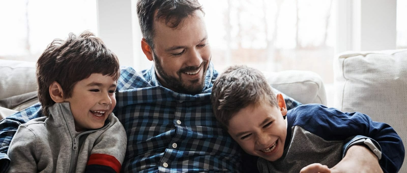 Pai sentado no sofá com duas crianças, os três estão rindo