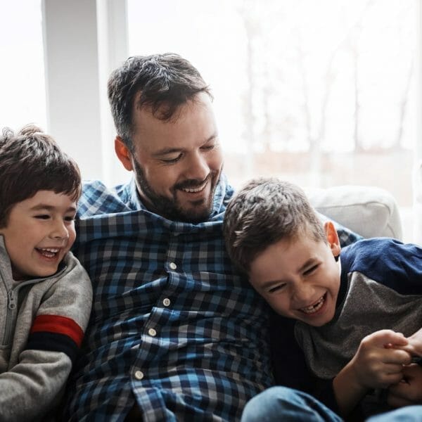 Pai sentado no sofá com duas crianças, os três estão rindo