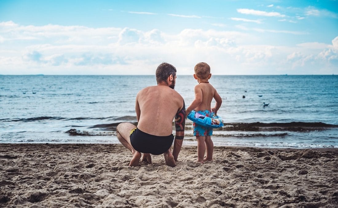 Pai e filho na praia. O pai está agachado na areia e o filho está ao lado, usando uma boia. Ao fundo, aparece o mar e um céu azul