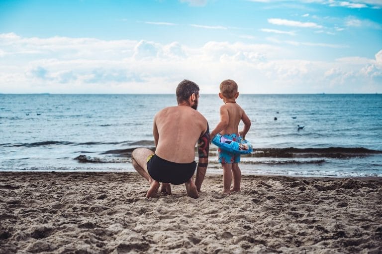 Pai e filho na praia. O pai está agachado na areia e o filho está ao lado, usando uma boia. Ao fundo, aparece o mar e um céu azul