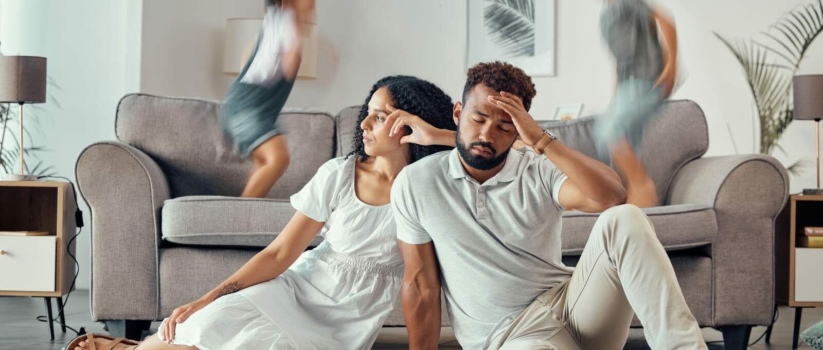 pais emburrados e sentados no chão à frente da imagem. em segundo plano, crianças pulando aceleradas em cima do sofá da sala