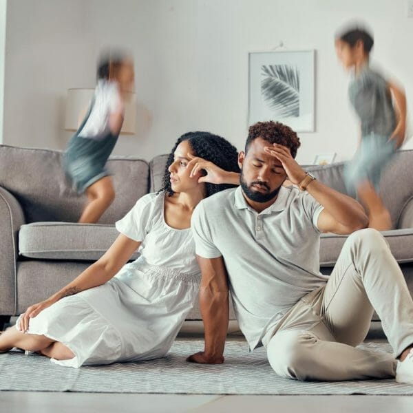 pais emburrados e sentados no chão à frente da imagem. em segundo plano, crianças pulando aceleradas em cima do sofá da sala